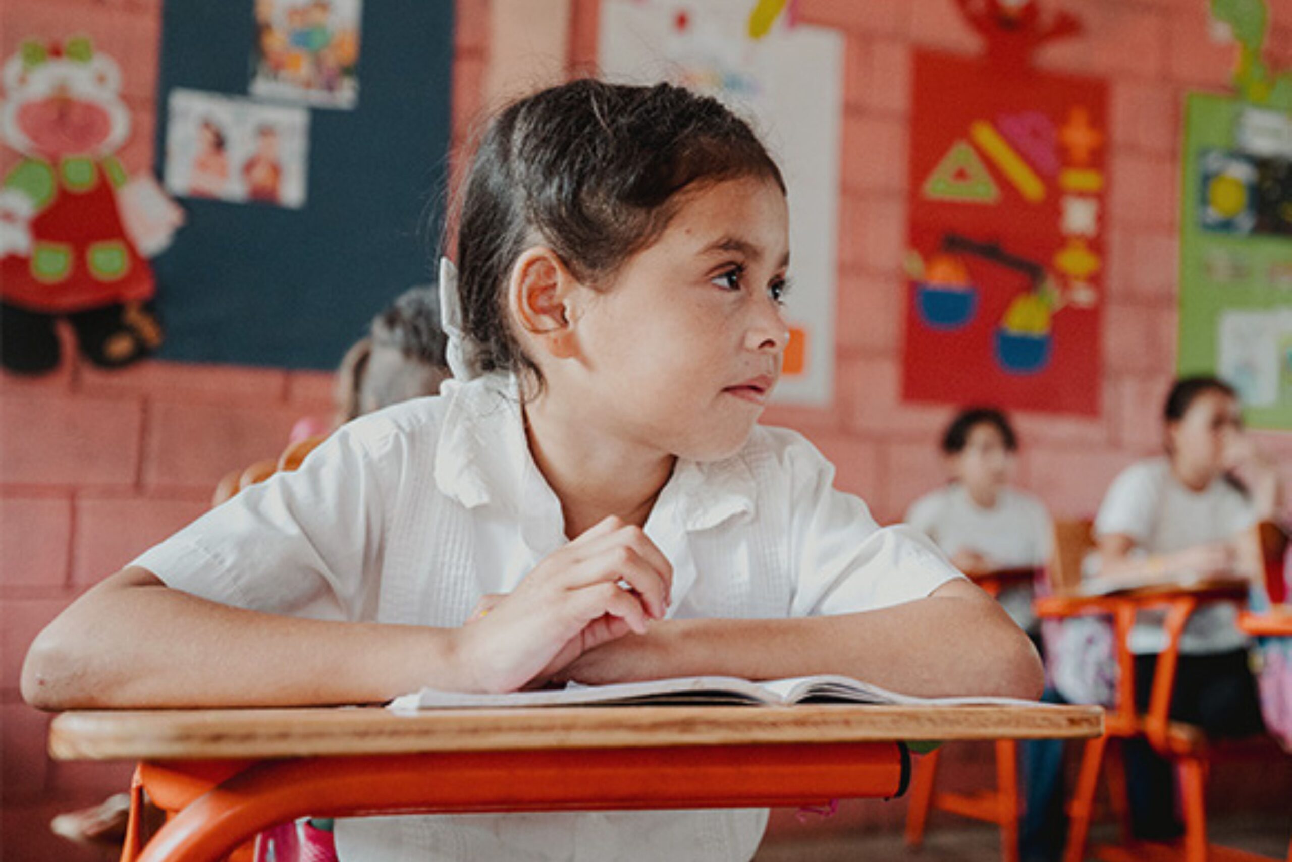 A young child is seated at a desk in a classroom with red walls.