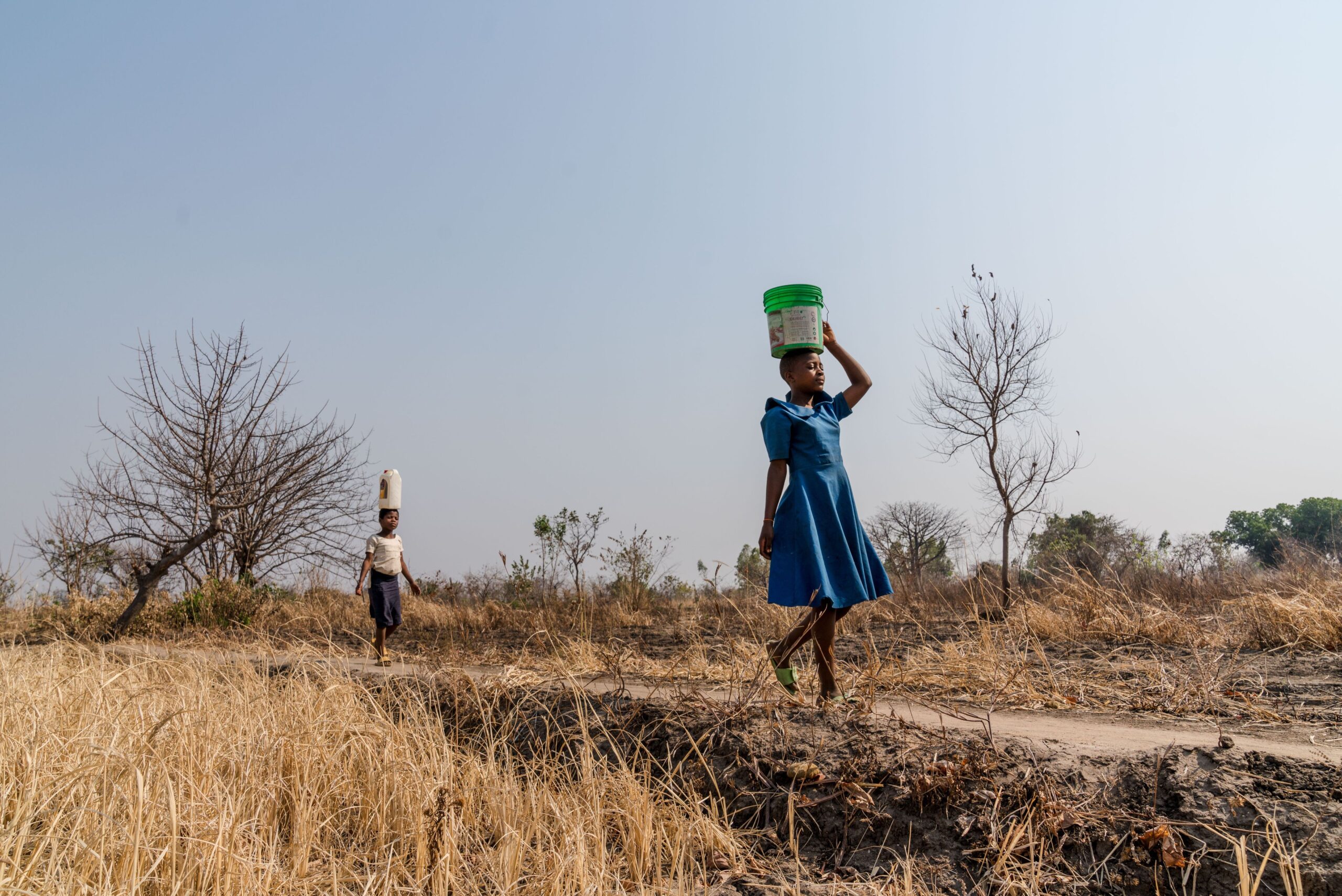 A young girl wearing a blue dress walks in a dry field, balancing a green bucket on her head.
