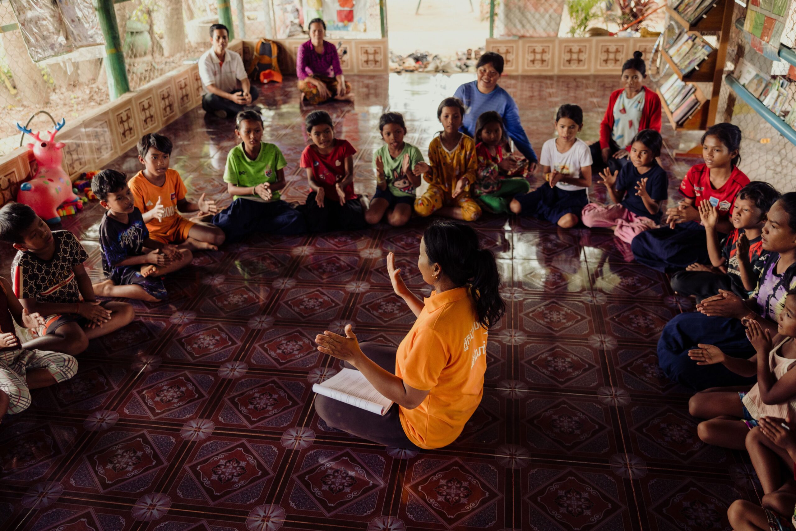 A group of children sit in a circle on the floor, listening to a World Vision staff member speaking in the middle.