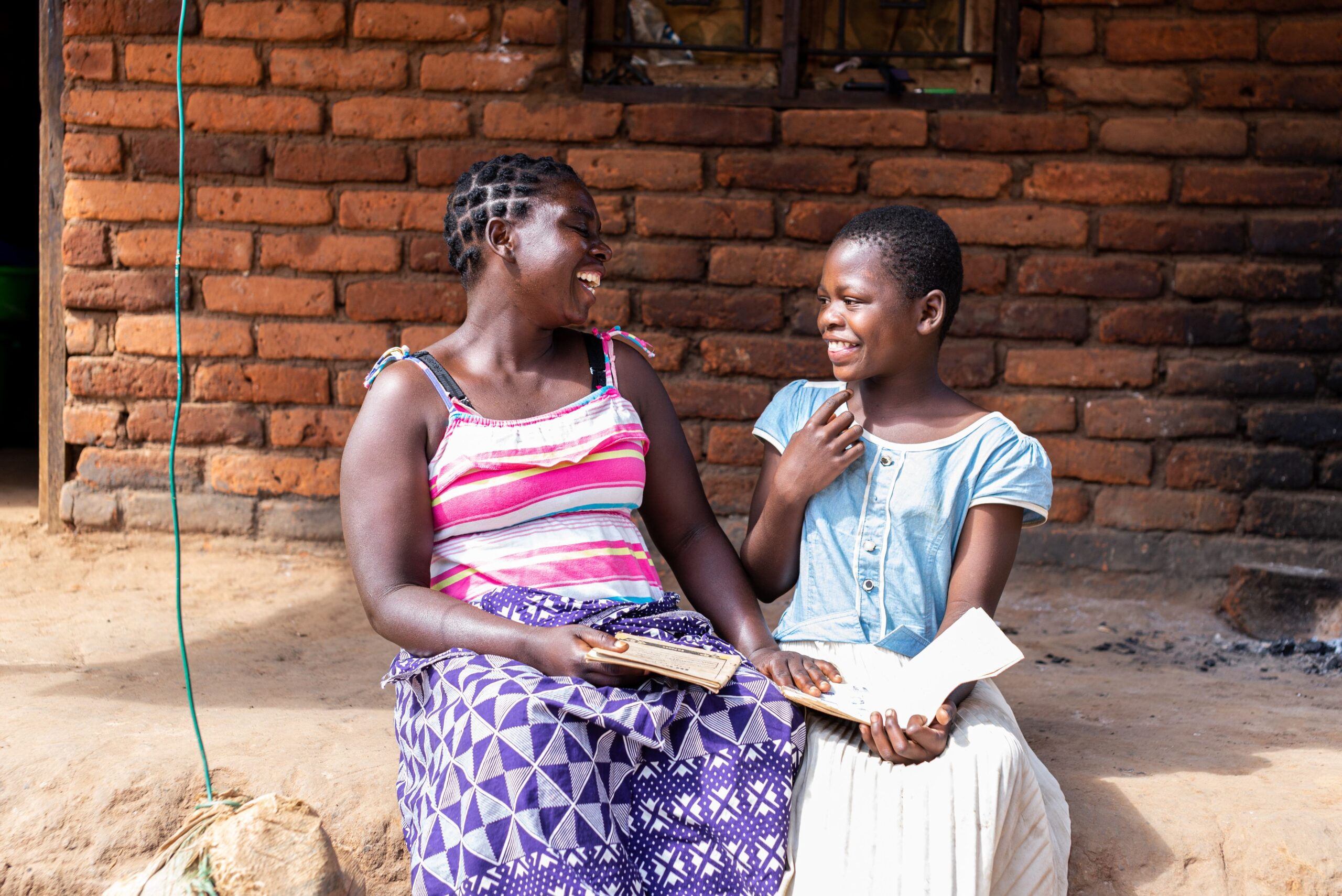 A mother and daughter laugh as they sit on the ground in front of a brick wall.
